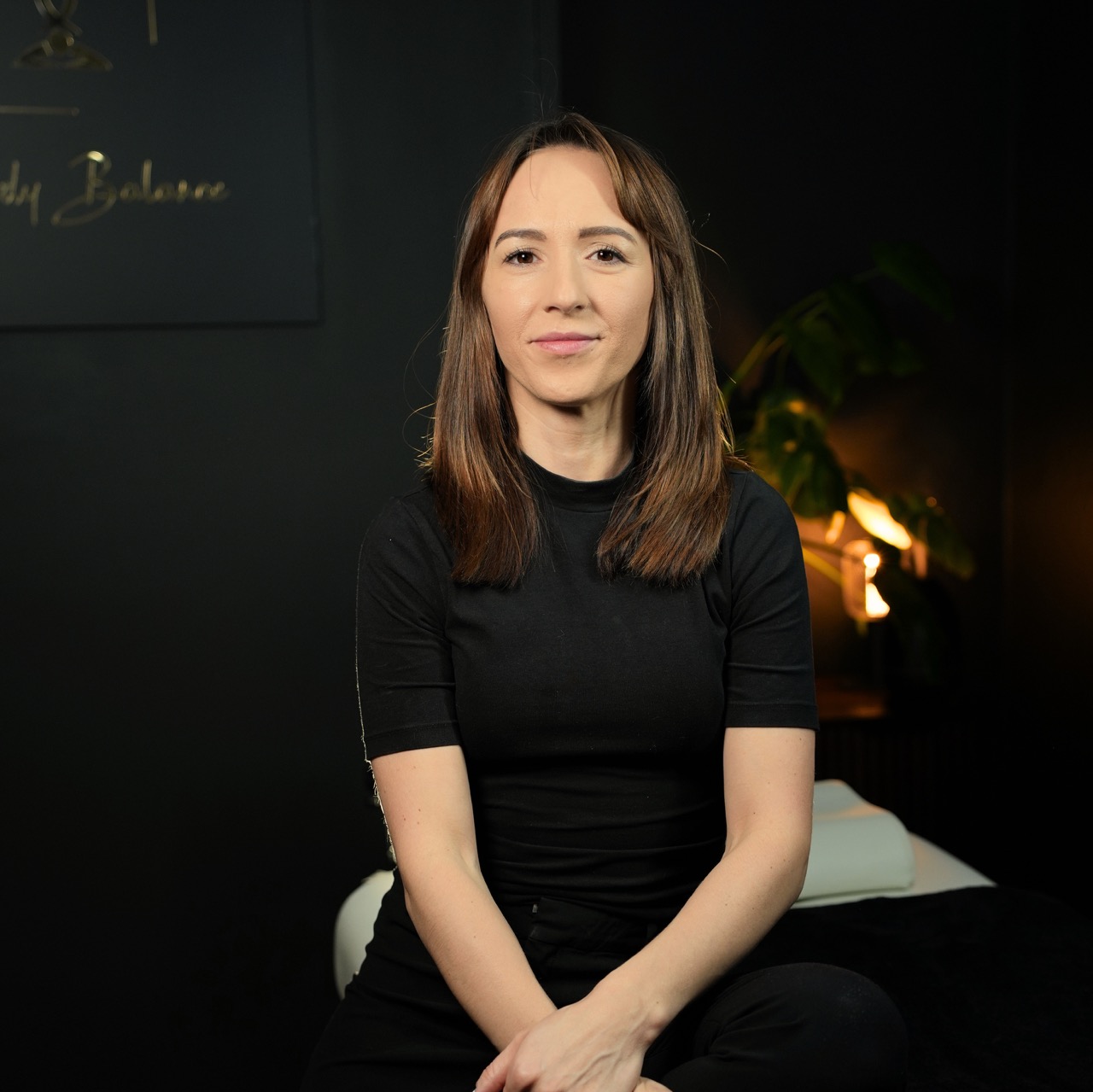 A female massage therapist with straight, shoulder-length brown hair sits facing the camera in a softly lit, dark treatment room. She wears a fitted black short-sleeve top and has a calm, welcoming expression. The background is dim, with a blurred massage table, a plant, and warm ambient lighting that creates a relaxing, spa-like atmosphere.